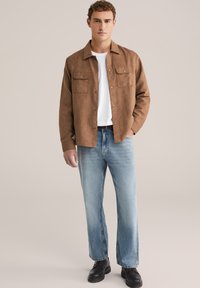 Bruine suède jas met twee borstzakken, een wit t-shirt eronder, lichtblauwe denim jeans en zwarte veterschoenen, weergegeven op een effen achtergrond.