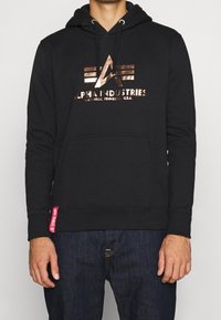 Sudadera negra con capucha, bolsillo tipo canguro, logo metálico en bronce con detalles geométricos y etiqueta roja en el dobladillo. Material de mezcla de algodón.