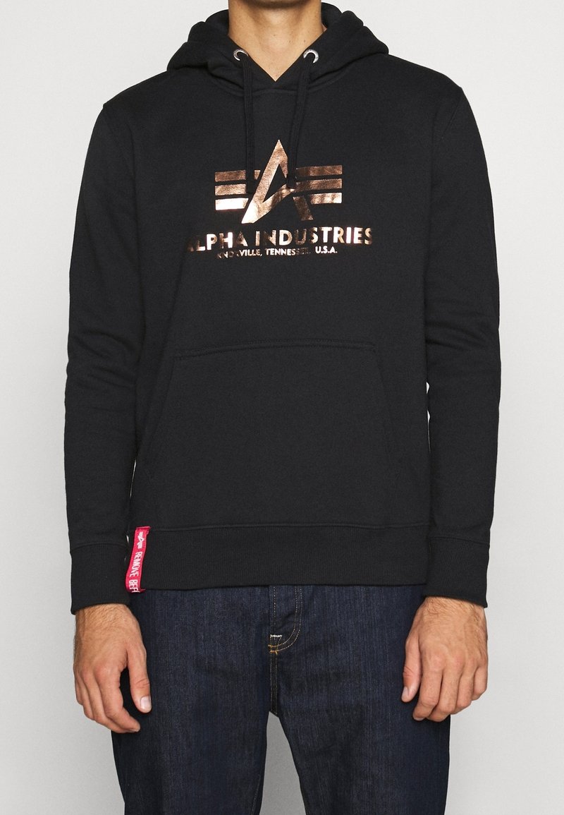 Sudadera negra con capucha, bolsillo tipo canguro, logo metálico en bronce con detalles geométricos y etiqueta roja en el dobladillo. Material de mezcla de algodón.