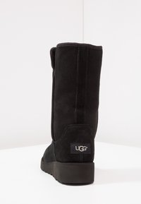 Czarna zamszowa kozaczka za kolano o zaokrąglonym nosku, płaskiej podeszwie i naszywce z logo UGG. Miękka faktura i prosty design z przeszyciami z tyłu.