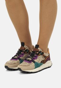 Scarpe sportive multicolori con una parte superiore maculata beige, dettagli viola e verdi, e una suola testurizzata. Presentano un mix di materiali, inclusa la pelle scamosciata.