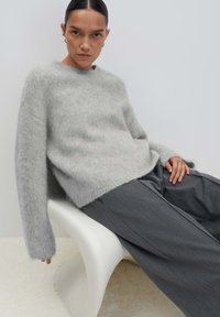 Vrouw met donker haar dat naar achteren is getrokken, draagt een fuzzy lichtgrijze sweater en wijd donkere grijze broek, zittend op een moderne witte stoel tegen een effen achtergrond.