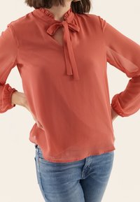 Blusa corallo realizzata in tessuto trasparente, con colletto arricciato, fiocco annodato e maniche a sbuffo, indossata su jeans blu.
