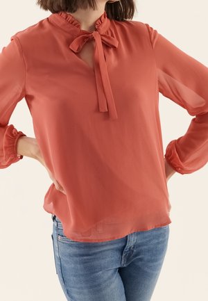 Blouse corail en tissu transparent avec un col volanté, un nœud noué et des manches bouffantes, portée sur un jean bleu.