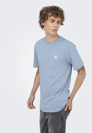 Joven con pelo rizado que lleva una camiseta azul claro "only and sons" y vaqueros negros desgastados, mirando hacia la izquierda con tatuajes visibles en ambos brazos.