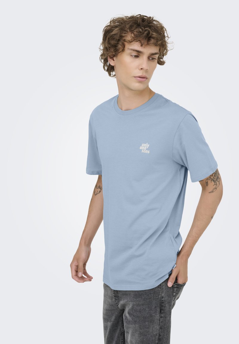 Joven con pelo rizado que lleva una camiseta azul claro "only and sons" y vaqueros negros desgastados, mirando hacia la izquierda con tatuajes visibles en ambos brazos.