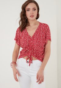 Rød blomsterbluse med V-hals, korte ærmer og sammensnoet detalje ved taljen. Sammen med hvide jeans. Glat stofstruktur.