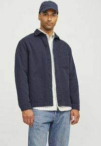 Chaqueta de botón azul marino con un bolsillo en el pecho, hecha de tela de algodón. Superpuesta sobre una camisa de rayas, combinada con unos vaqueros de mezclilla claros.