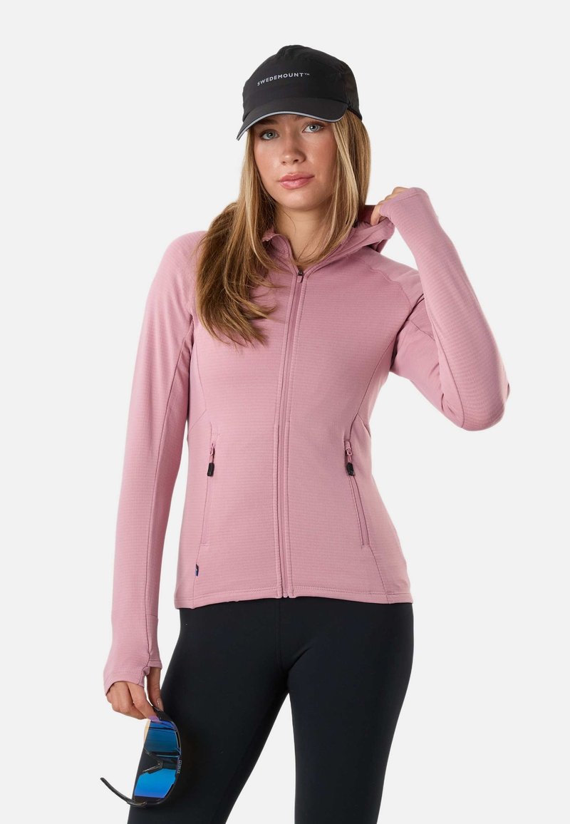Frau trägt eine schwarze Kappe, eine pinke Zip-Up-Jacke mit Daumenlöchern und schwarze Leggings und hält eine Sonnenbrille mit blauen Gläsern in einer Hand.