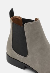 Botín Chelsea de ante gris con panel lateral elástico negro, asa negra y suela negra, mostrado en primer plano.