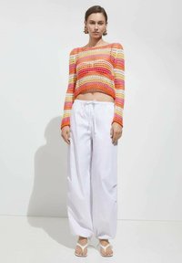 Haken-stijl top met horizontale strepen in oranje, roze en geel; witte wijde broek; platte witte sandalen; gladde textuur.