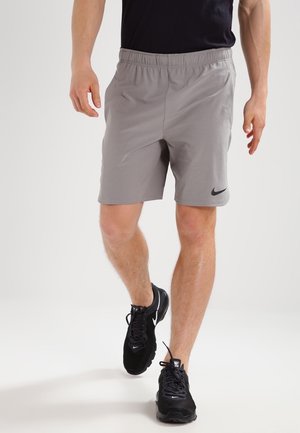 Mann, der hellgraue Nike-Shorts und schwarze Nike-Laufschuhe trägt, vor einem schlichten weißen Hintergrund geht.