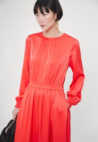 Rotes satinlangärmeliges Kleid mit rundem Ausschnitt, gerafften Details, elastischem Taillenbund und elastischen Manschetten. Kombiniert mit einer schwarzen Handtasche.