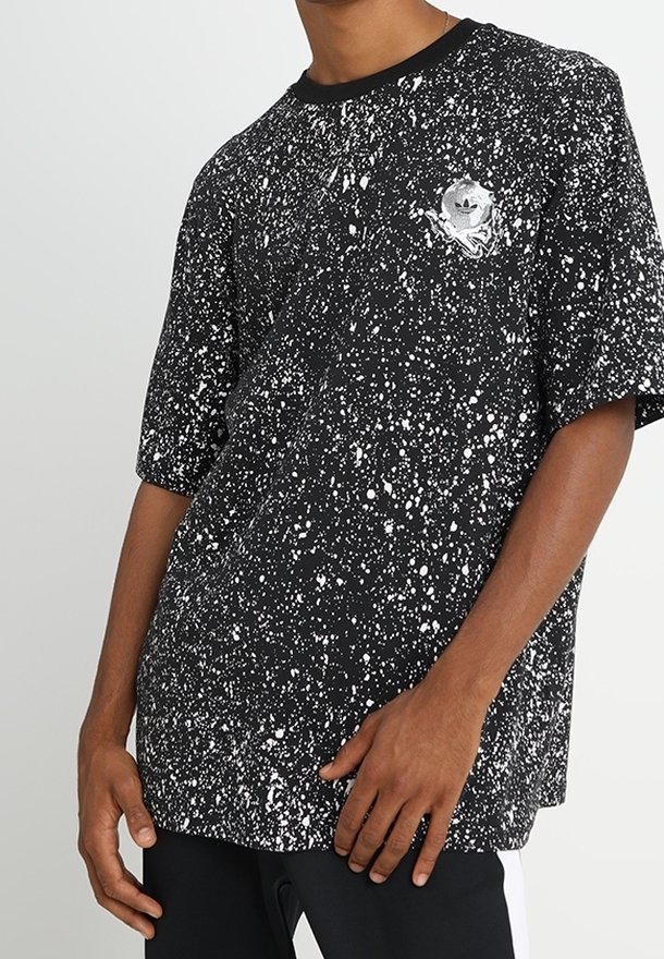 Černé tričko s bílým stříkavým vzorem a malým stříbrným logem Adidas na hrudi, nošené osobou s tmavou pletí.