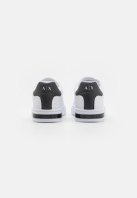 White sneakers with textured side panels and a black heel tab. Features a black stripe on the sole and a logo on the back.