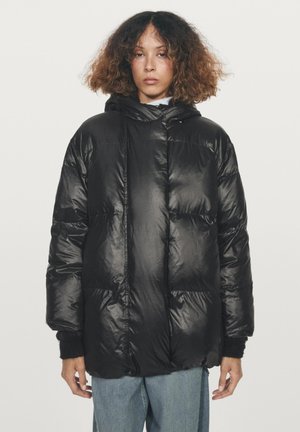 Kvinna med lockigt hår klädd i en svart puffjacka och ljusblå jeans som står mot en helt vit bakgrund.