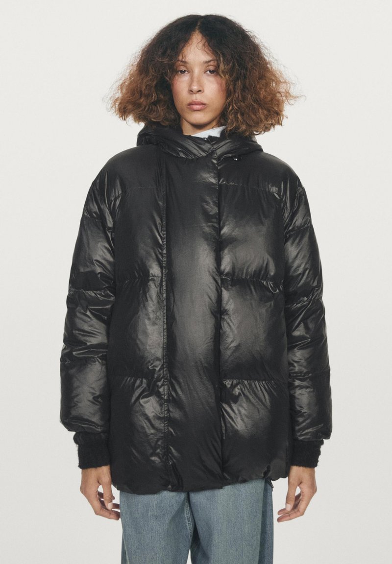 Žena s kudrnatými vlasy, oblečená v černé prošívané bundě a světlemodrých džínech, stojící na jednobarevném bílém pozadí.