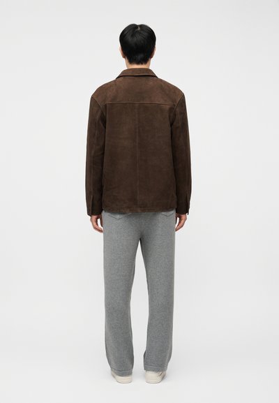 Άνδρας που είναι γυρισμένος με την πλάτη, φοράει σκούρο καφέ σακουλέ τζάκετ, ανοιχτό γκρι παντελόνι και λευκά αθλητικά παπούτσια σε ένα απλό λευκό φόντο.