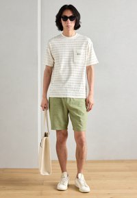 Beige tekstureret t-shirt med en brystlomme, parret med grønne shorts, hvide sneakers og en beige tote bag. Model iført sorte solbriller.
