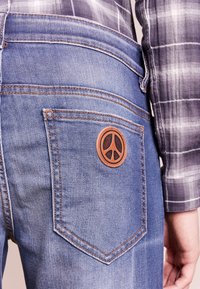 Modré džíny vyrobené z denimu s koženou nášivkou symbolu míru na zadní kapse, s kontrastním béžovým stehem a hladkou texturou.