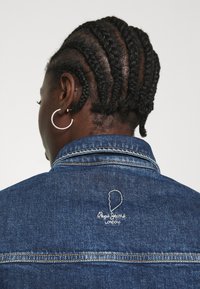 Jeansjacke in Dunkelblau mit klassischem Kragen und vernickelter Hardware. Besitzt ein weißes, gesticktes Logo auf der Rückseite und einen strukturierten Stoff.