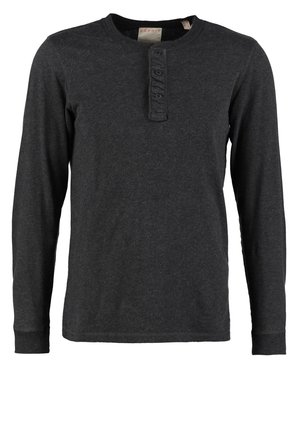 Beige kabelstrikket cardigan over en sort henley-shirt, kombineret med mørkeblå jeans og sorte ankelstøvler. Afslappet, tekstureret stof med knapper.