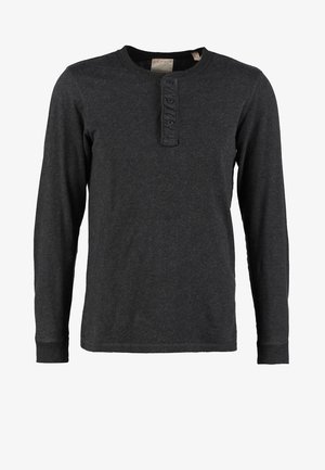 Beżowy kardigan z grubym splotem na czarnej koszulce typu henley, zestawiony z ciemnoniebieskimi dżinsami i czarnymi botkami za kostkę. Swobodny styl, teksturowana tkanina z guzikami.