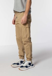 Khaki cargo hlače s elastičnim manžetama, bočnim džepovima i opuštenim krojem, uparene s tamnoplavo-bijelim tenisicama s teksturiranim detaljima.