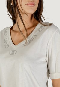 T-shirt beige chiaro a scollo a V realizzato in tessuto morbido. Presenta un delicato motivo con logo in cristalli argentati lungo il collo e le spalle. Maniche corte.