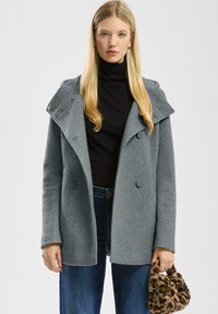Femme portant un manteau en laine gris sur un col roulé noir et un jean bleu, tenant à la main gauche un petit sac à main en fausse fourrure imprimé léopard.
