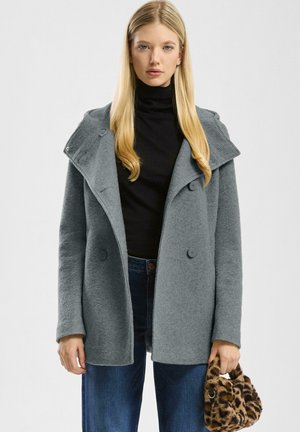 Femme portant un manteau en laine gris sur un col roulé noir et un jean bleu, tenant à la main gauche un petit sac à main en fausse fourrure imprimé léopard.