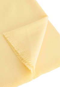 Žlutá tkanina s hladkou texturou, zdobená třepeným okrajem. Tkanina je částečně složená, což ukazuje na její barvu a povrchovou úpravu.