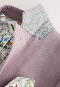 Indumento viola con colletto in feltro grigio; l'interno presenta una fodera floreale con colori pastello e motivi curvi, conferendo un aspetto testurizzato.