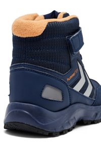 Bota azul marino con parte superior de tela texturizada, cuello de forro polar naranja y detalles en gris. Presenta una suela resistente y una correa de Velcro para el cierre.