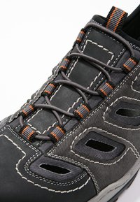 Chaussure de randonnée noire et grise avec une tige texturée, des accents de lacets orange, plusieurs découpes de ventilation et des détails de couture blanche.