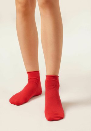 Legs wearing bright red ankle socks against a neutral background, positioned with one foot slightly forward and toes pointed down.