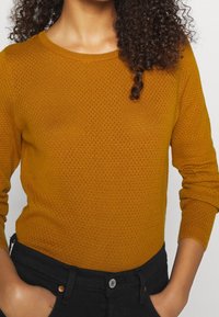 Pull tricoté couleur moutarde avec un col rond, des manches longues et un motif texturé. Associé à un pantalon noir taille haute.