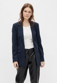 Navy blue blazer with a tailored design, featuring notched lapels, side pockets, and smooth fabric. Worn over a white ribbed top and black pants.