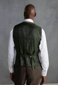 Gilet vert à motifs avec une texture lisse, détail de sangle au dos, porté sur une chemise blanche, associé à un pantalon brun.