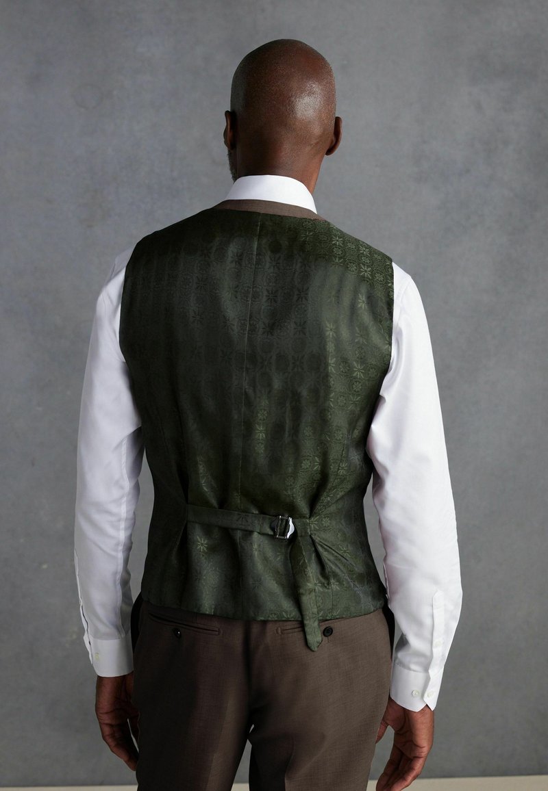 Gilet vert à motifs avec une texture lisse, détail de sangle au dos, porté sur une chemise blanche, associé à un pantalon brun.