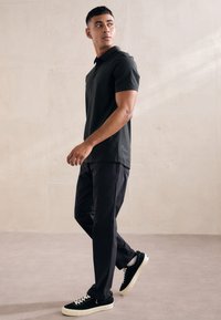 Cămașă polo neagră, pantaloni închiși la culoare și adidași negri cu șireturi albe. Modelul stă în fața unui fundal deschis, evidențiind o ținută casual.