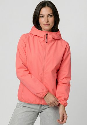Frau trägt korallfarbenen Kapuzenjacke mit Reißverschluss, zieht Ärmel hoch, steht vor schlichtem hellem Hintergrund.