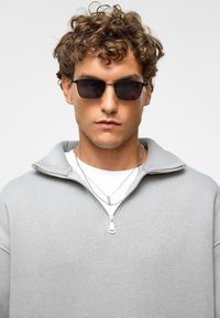 Grijze sweatshirt met rits, gemaakt van zacht materiaal, gecombineerd met een paar donkere zonnebril. Gelaagde zilveren kettingen voegen detail toe aan de outfit.