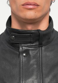 Black leather jacket with a high collar, featuring snap buttons and a textured finish. Visible stitching along the front adds detail.