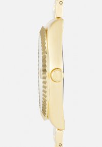 Montre analogique en or avec un bezel texturé et rainuré, un boîtier circulaire et une couronne ronde, accompagnée d'un bracelet en métal lisse et d'une finition polie.