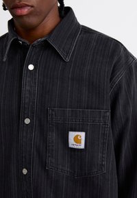 Chemise à boutons rayée noire en denim avec une poche poitrine et un patch Carhartt orange cousu. Boutons-pression argentés visibles.
