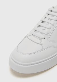 Bijela kožna tenisica s glatkom teksturom, okruglim vrhom i šivenim detaljima. Ima bijele vezice i debeli gumeni potplat.