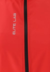 Giacca rossa con zip frontale e rifiniture nere, realizzata in tessuto liscio. Il logo "ELITE LAB" è stampato in argento sul lato sinistro.