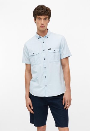 Jeune homme aux cheveux bruns bouclés portant une chemise bleu clair à manches courtes avec poches poitrine et un short bleu marine foncé.