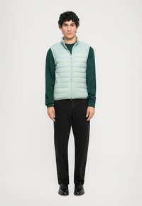 Giovane in vita un gilet trapuntato azzurro su una camicia a maniche lunghe verde scuro, pantaloni neri e scarpe nere, in piedi davanti a uno sfondo bianco.